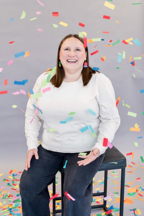 Julie Thoman sitting in a chair, smiling and looking up as rainbow confetti falls over her.